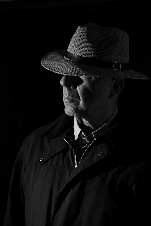 Man in a fedora and trench coat lit in film-noir shadow